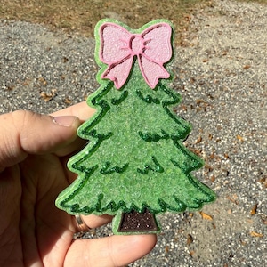 May include: A glittery green Christmas tree ornament with a pink bow at the top. The tree has a brown trunk and a green outline. The bow has a pink outline. The ornament is held in a hand.