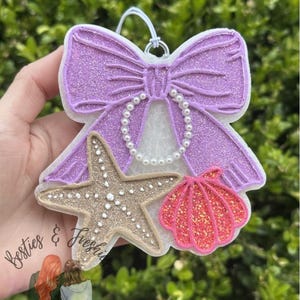 K&ouml;nnte beinhalten: Ein h&auml;ngendes Lufterfrischer-Ornament mit einer gro&szlig;en, glitzernden lila Schleife, die mit einer Kette aus wei&szlig;en Kunstperlen verziert ist. Unter der Schleife sind ein strukturierter beiger Seestern und eine leuchtend rosa glitzernde Muschel befestigt.