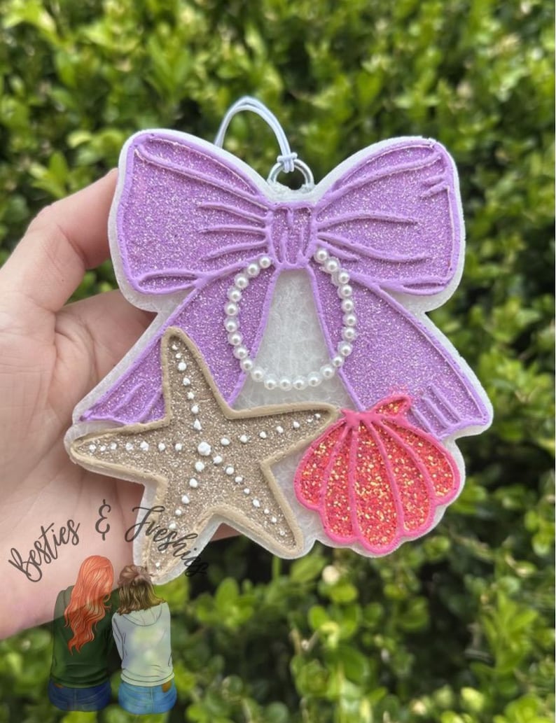 K&ouml;nnte beinhalten: Ein h&auml;ngendes Lufterfrischer-Ornament mit einer gro&szlig;en, glitzernden lila Schleife, die mit einer Kette aus wei&szlig;en Kunstperlen verziert ist. Unter der Schleife sind ein strukturierter beiger Seestern und eine leuchtend rosa glitzernde Muschel befestigt.