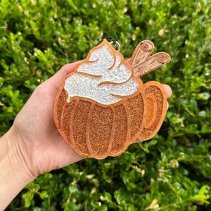 May include: A glittery orange pumpkin-shaped ornament with a white, sparkly whipped cream topping and two cinnamon sticks. The ornament has a black loop for hanging. The design is reminiscent of a pumpkin spice latte.