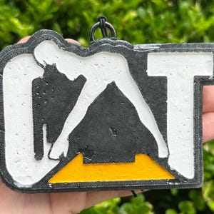 May include: A rectangular black, white, and yellow 3D printed keyring featuring the letters CAT in a bold, block font. The design includes a black triangular shape at the bottom centre, a white background, and a black border with a small metal ring.