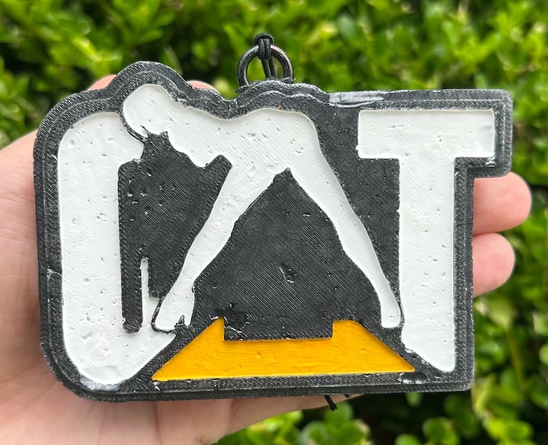 May include: A rectangular black, white, and yellow 3D printed keyring featuring the letters CAT in a bold, block font. The design includes a black triangular shape at the bottom centre, a white background, and a black border with a small metal ring.