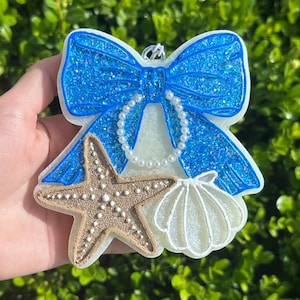 K&ouml;nnte beinhalten: Ein dekoratives Ornament mit einer gro&szlig;en, glitzernden blauen Schleife und einer wei&szlig;en, perlenartigen Schlaufe in der Mitte. Unter der Schleife sind ein strukturierter beiger Seestern und eine wei&szlig;e Muschel auf der wei&szlig;en Basis befestigt.