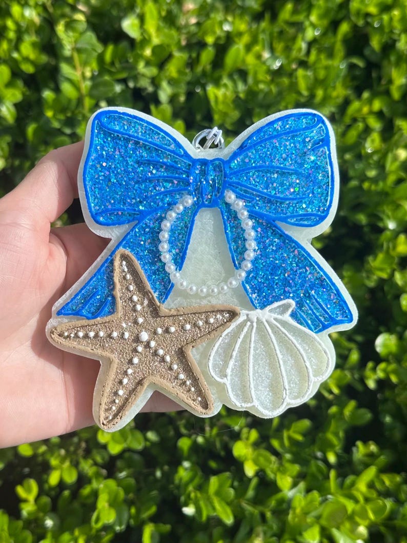 K&ouml;nnte beinhalten: Ein dekoratives Ornament mit einer gro&szlig;en, glitzernden blauen Schleife und einer wei&szlig;en, perlenartigen Schlaufe in der Mitte. Unter der Schleife sind ein strukturierter beiger Seestern und eine wei&szlig;e Muschel auf der wei&szlig;en Basis befestigt.