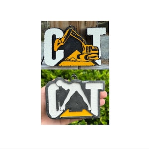 May include: Two rectangular hanging ornaments featuring the letters CAT. The top ornament displays a yellow excavator graphic integrated into the letter A. The bottom ornament features a white silhouette of a person in a pose within the letter A.