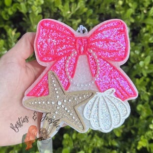 K&ouml;nnte beinhalten: Ein dekorativer Lufterfrischer in Form einer gro&szlig;en, glitzernden, pinkfarbenen Schleife mit wei&szlig;en perlenartigen Akzenten, einem beigen, strukturierten Seestern und einer wei&szlig;en, irisierenden Muschel mit kleiner Metall&ouml;se zum Aufh&auml;ngen.