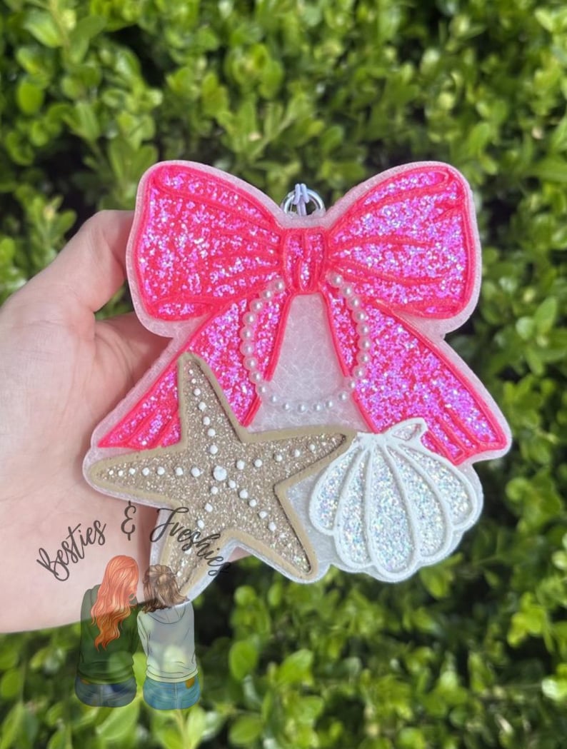 K&ouml;nnte beinhalten: Ein dekorativer Lufterfrischer in Form einer gro&szlig;en, glitzernden, pinkfarbenen Schleife mit wei&szlig;en perlenartigen Akzenten, einem beigen, strukturierten Seestern und einer wei&szlig;en, irisierenden Muschel mit kleiner Metall&ouml;se zum Aufh&auml;ngen.