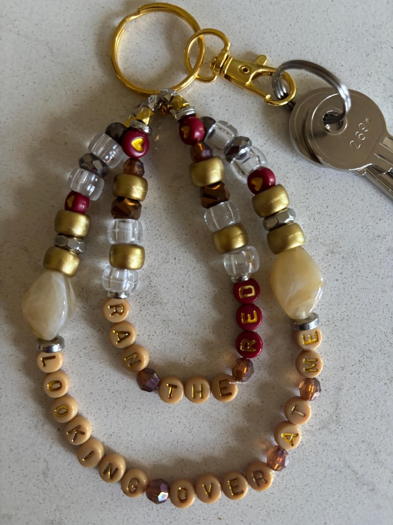 May include: A gold key chain with a beaded charm that reads "Looking over at the same time". The beads are brown, gold, clear, and red. The charm is made of beige and brown beads.