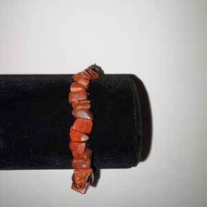 May include: A bracelet made of reddish-brown, irregularly shaped stones. The stones have a glittery, speckled appearance. The bracelet is displayed against a black background, highlighting its color and texture. The jewelry piece is a simple, elegant accessory.