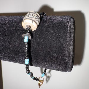 May include: A beaded bracelet with a cylindrical, off-white bead with a black swirl design. The bracelet features black and turquoise beads, and a gold-coloured clasp. The bracelet is displayed on a black surface.