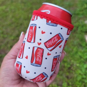 May include: A white and red Coca-Cola can cooler with a red rim, holding a Coca-Cola can. The cooler has a repeating pattern of Coca-Cola cans, red hearts, and black stars.