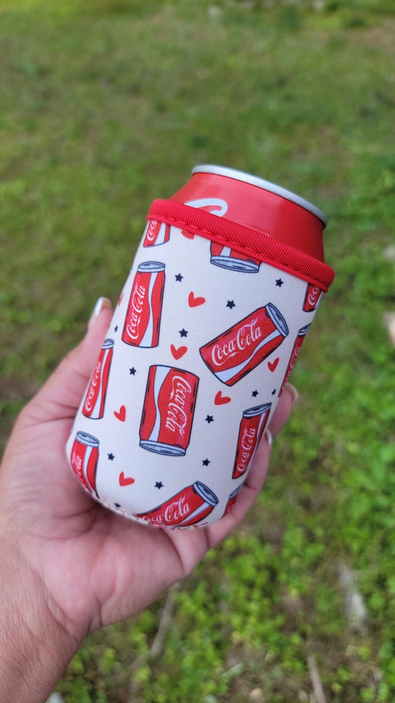 May include: A white and red Coca-Cola can cooler with a red rim, holding a Coca-Cola can. The cooler has a repeating pattern of Coca-Cola cans, red hearts, and black stars.