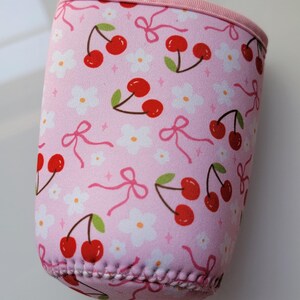 May include: Pink drinks sleeve featuring a pattern of red cherries, white flowers, and pink bows. The top and trim are pink. A round sticker with the text "Cute Gifts" is in the background.