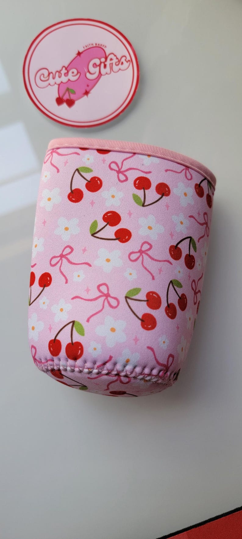 May include: Pink drinks sleeve featuring a pattern of red cherries, white flowers, and pink bows. The top and trim are pink. A round sticker with the text "Cute Gifts" is in the background.