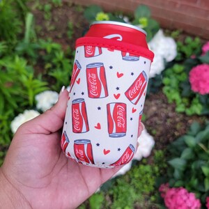 May include: A white and red Coca-Cola can cooler with a red rim. The cooler has a repeating pattern of Coca-Cola cans, red hearts, and blue stars. It is designed to keep drinks cool.