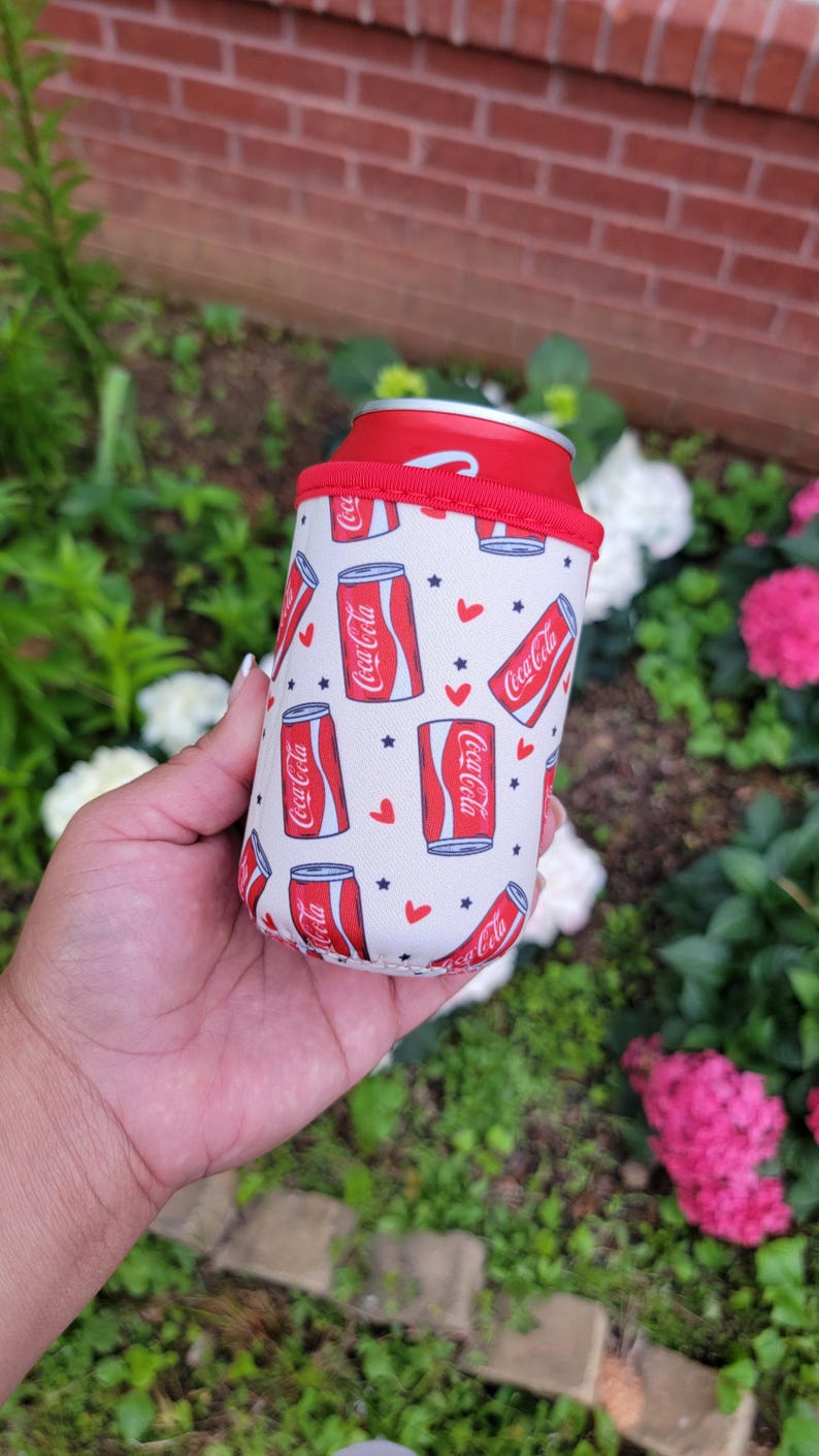 May include: A white and red Coca-Cola can cooler with a red rim. The cooler has a repeating pattern of Coca-Cola cans, red hearts, and blue stars. It is designed to keep drinks cool.
