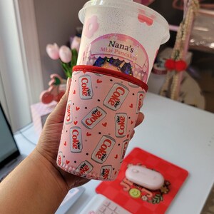 May include: A clear plastic cup with a straw, featuring a pink floral design and the text "Nana's Mini Pancakes." The cup is held in a pink Diet Coke can-themed cozy. A pink mouse and keyboard are also visible.