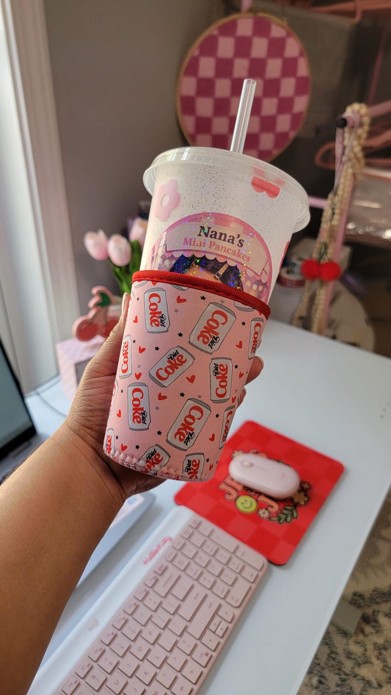 May include: A clear plastic cup with a straw, featuring a pink floral design and the text "Nana's Mini Pancakes." The cup is held in a pink Diet Coke can-themed cozy. A pink mouse and keyboard are also visible.