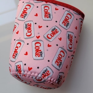 May include: Pink drinks can cooler with a repeating pattern of Diet Coke cans, hearts, and stars. The cooler has a red trim and is designed to keep drinks cold. A circular sticker with the text "Cute Gifts" is in the background.