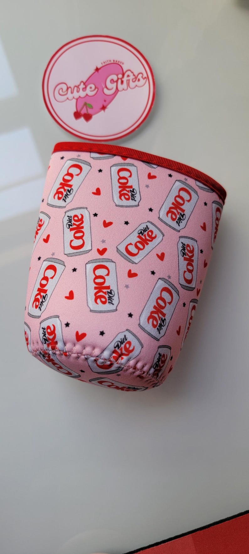 May include: Pink drinks can cooler with a repeating pattern of Diet Coke cans, hearts, and stars. The cooler has a red trim and is designed to keep drinks cold. A circular sticker with the text "Cute Gifts" is in the background.