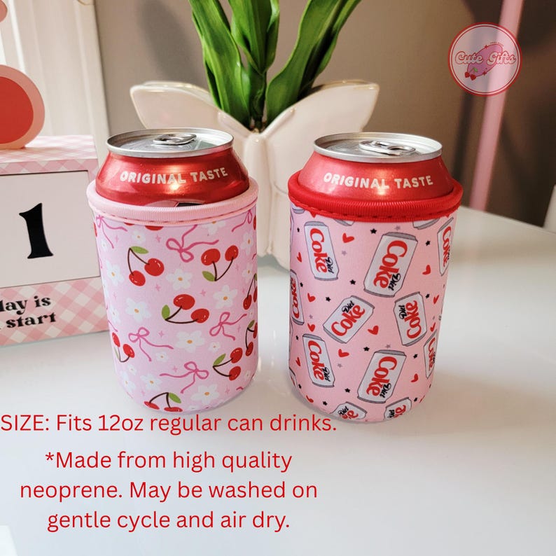 May include: Two pink can coolers with red rims, each holding a can. One cooler has a cherry pattern, the other has a Coke design. The text "ORIGINAL TASTE" is visible on the cans. The coolers fit 355ml regular can drinks.