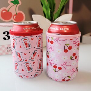 May include: Two pink can coolers with red trim, each holding a Diet Coke can. One cooler has a pattern of Diet Coke cans and hearts, the other has cherries and bows. The cans have "ORIGINAL TASTE" printed on the top.
