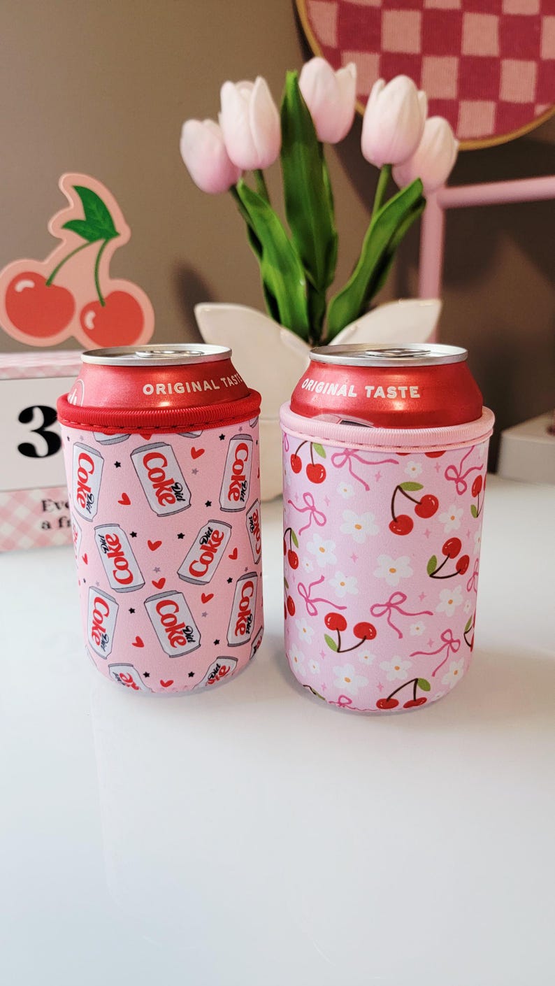 May include: Two pink can coolers with red trim, each holding a Diet Coke can. One cooler has a pattern of Diet Coke cans and hearts, the other has cherries and bows. The cans have "ORIGINAL TASTE" printed on the top.