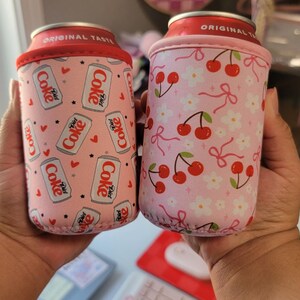 May include: Two pink can coolers. One has a pattern of Diet Coke cans, hearts, and stars. The other features a cherry and flower design. Both have a red rim and are designed to keep drinks cold.