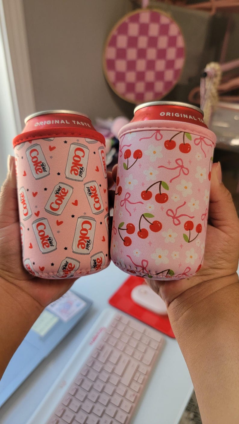 May include: Two pink can coolers. One has a pattern of Diet Coke cans, hearts, and stars. The other features a cherry and flower design. Both have a red rim and are designed to keep drinks cold.