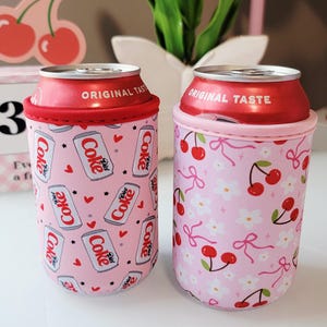 May include: Two drinks can coolers with red tops, each holding a Diet Coke can. One cooler is pink with Diet Coke cans and hearts, the other is pink with cherries and bows. A sticker with the text "Cute Gifts" is visible.
