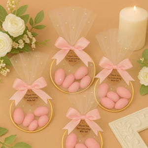 Può includere: Quattro bomboniere decorative con mandorle rosa zuccherate all'interno di cerchi dorati. Ogni bomboniera è adornata con un fiocco rosa, un tessuto trasparente e un'etichetta con la scritta "Thank You For Calimasting With Us!". Una candela bianca e fiori sullo sfondo.