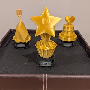 May include: Three gold-colored 3D printed trophies on a brown surface. The trophies are shaped like a star, a cupcake, and a heart-shaped cake. The star trophy has the text "STAR BAKER 1st PLACE" on the base. The cupcake trophy has the text "CUPCAKE CHAMPION" on the base. The heart-shaped cake trophy has the text "BEST DESSERT" on the base.