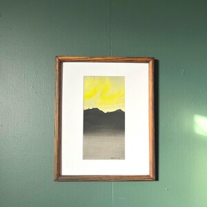 May include: A framed watercolor landscape painting. The artwork features a gradient of colors, with a bright yellow sky above a dark gray mountain range and a gray foreground. The frame is made of wood.