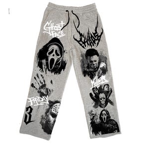 May include: Light grey sweatpants featuring black and white graphic prints of horror movie characters and motifs. The designs include the text Ghost Face, Chaos, Friday, 3, and Killer, alongside illustrations of masked figures and a handprint.
