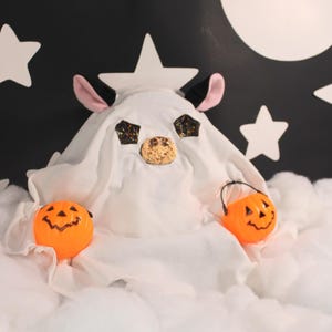 May include: A Halloween-themed display featuring a white ghost costume with pink ears, black horns, and glittery eyes. Two orange jack-o'-lantern buckets and a cookie nose sit on a bed of fluffy white clouds against a black backdrop with white stars and a moon.