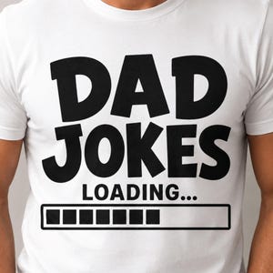 Peut inclure: Un t-shirt blanc à manches courtes avec le texte noir en gras DAD JOKES LOADING... suivi d'un graphique de barre de progression rectangulaire noir avec huit carrés remplis, centré sur le devant du vêtement.