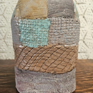 以下が含まれることがあります： パッチワークデザインの手作りセラミック花瓶。花瓶は、茶色、グレー、ティール色のテクスチャード加工された正方形が特徴です。小さな丸いティール色のノブが上にあります。花瓶は木製の表面に展示されています。