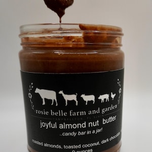 May include: A glass jar filled with dark brown almond butter, with a spoon dripping the butter. The label reads "rosie belle farm and garden" and "joyful almond nut butter...candy bar in a jar!"  The label also lists the ingredients as "roasted almonds, toasted coconut, dark chocolate" and the size as "9 ounces".