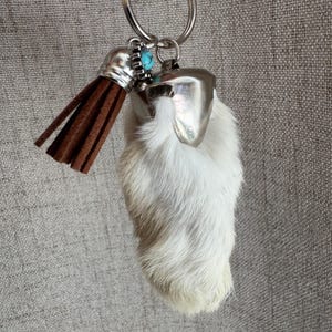 May include: A keychain featuring a white fur foot with a silver-colored metal accent. A brown tassel and a turquoise-colored stone add detail. The keychain is attached to a silver-colored metal ring.