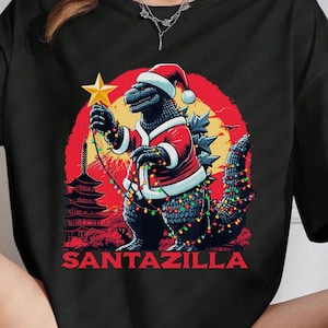 Può includere: Maglietta nera con una grafica di Godzilla vestito da Babbo Natale che tiene una stella e luci di Natale. Il testo "Santazilla" è stampato sotto la grafica.