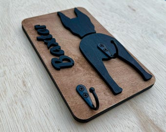 May include: Wooden dog lead hook with the name "Baxter" engraved on it. The hook is shaped like a dog's rear end and has a black finish.