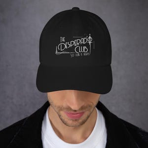 May include: A black baseball cap featuring white embroidered text that reads The Desperado Club So Fun It Hurts, with a stylized sword graphic integrated into the lettering on the front panel of the hat, which has a curved brim and structured crown.