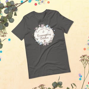 May include: A dark gray short-sleeve t-shirt featuring a circular floral wreath graphic. Inside the wreath, the text reads Unity, Family, Support & Kneecapping Bitches in a dark brown script and sans-serif font, centered on the chest area.