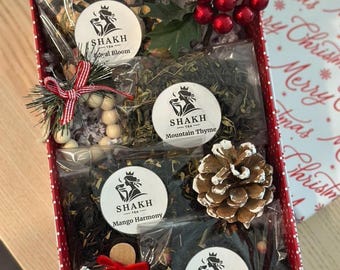 May include: A red and white polka dot gift box filled with tea packets, decorative elements, and a wooden ornament. The tea packets are labeled "Royal Bloom", "Mountain Thyme", "Mango Harmony", and "Berry Bloom". The box is decorated with holly and berries.