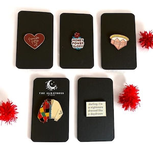May include: A collection of enamel pins on black cards. The pins feature various designs, including a heart with text, a rose with the words "Money Power Glory," and a sweater. One pin reads "darling, I'm a nightmare dressed like a daydream."