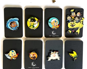 以下が含まれることがあります： 黒い長方形のカードに、漫画のキャラクターが描かれた8つのエナメルピンのコレクション。ピンは、青、黄、オレンジなど、さまざまな鮮やかな色でキャラクターを描いています。一部のカードには「THE ALBATROSS SHOP」の文字が入っています。