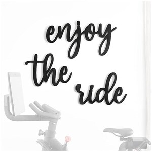 Könnte beinhalten: Schwarze Holz-Wanddekoration mit den Worten "enjoy the ride" in kursivschrift. Die Dekoration ist an einer weißen Wand über einem Heimtrainer angebracht.