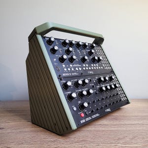 May include: A black and olive green synthesizer with numerous knobs and buttons. The synthesizer has a handle and is angled on a wooden surface. The text "Moog" is visible on the front panel, along with the model names "Mother-32" and "DFAM".