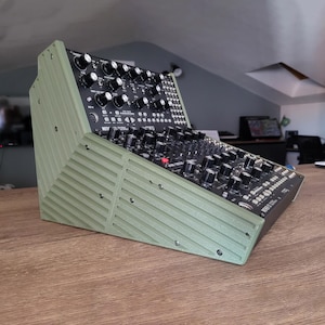 Puede incluir: Un sintetizador modular con un marco verde acanalado. El sintetizador tiene numerosas perillas y botones negros. El marco está en ángulo y el dispositivo se encuentra sobre una superficie de madera. El sintetizador es de la marca Moog.