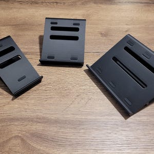 Puede incluir: Tres soportes para teléfono negros hechos de plástico. Los soportes están diseñados para sostener un teléfono en ángulo para verlo.